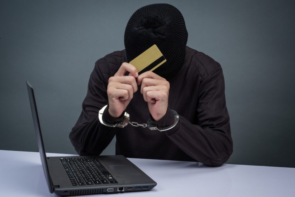 Cybercriminal in handcuffs holding a credit card near a laptop, representing identity theft and cybersecurity threats.