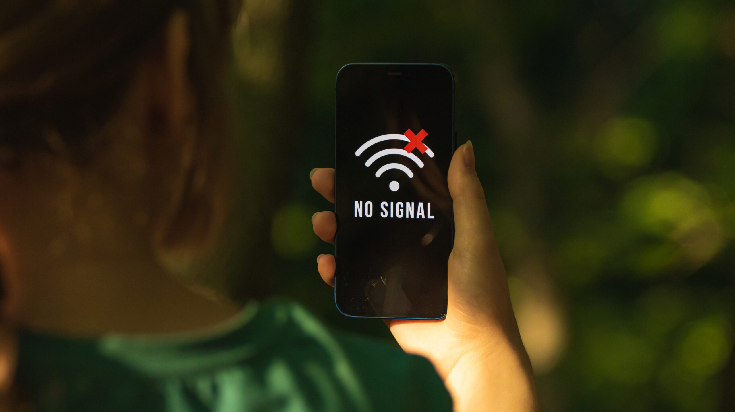 A woman holding a smartphone with a 'No Signal' screen in a forest, illustrating the challenges of communication failure and the importance of alternative methods during crises.