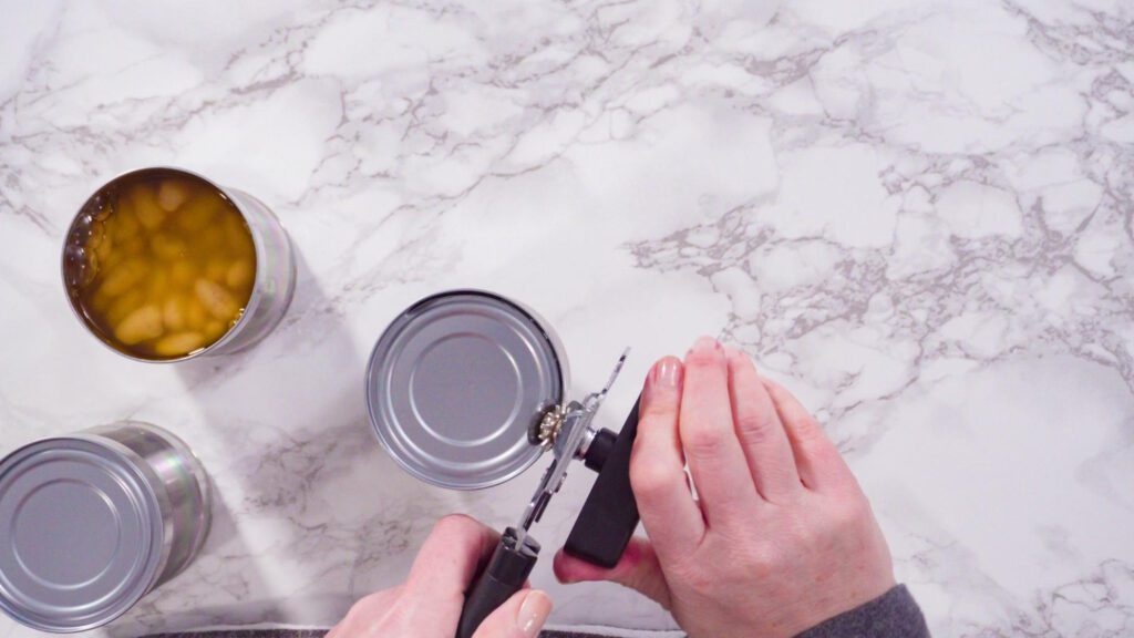 Hands using a manual can opener to open a tin can on a marble countertop, with an open can of food visible nearby.
