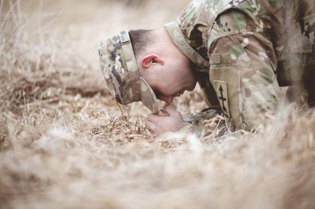 A fatigued soldier in camouflage uniform kneels in a dry field, hands clenched, head down, battling exhaustion and stress—symbolizing the mental fight for survival.