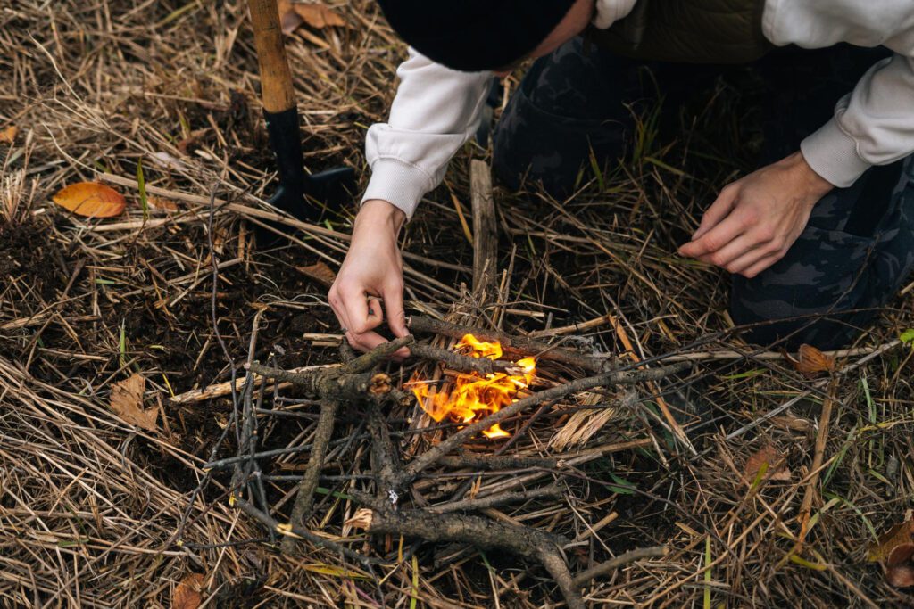A survivalist crouches in the wilderness, carefully tending to a small fire built from natural materials, demonstrating survival without gear in an emergency.
