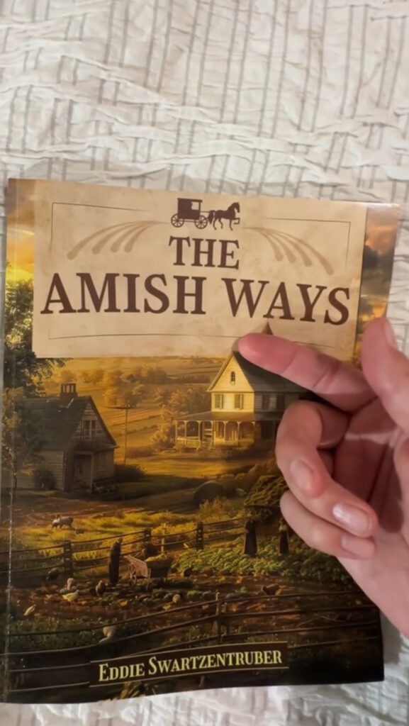 Close-up of The Amish Ways book by Eddie Swartzentruber, with a hand pointing at the title. The book cover features a traditional Amish farm scene, symbolizing self-sufficiency and survival skills.
