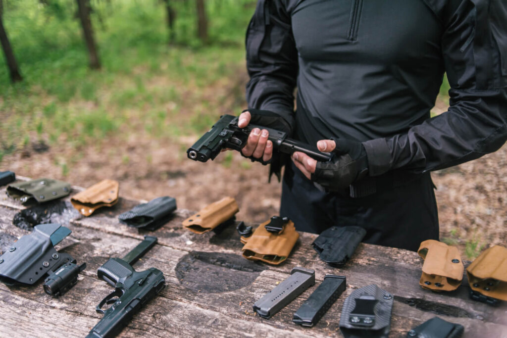 Man clearing handgun malfunction during outdoor survival training, surrounded by gear and spare magazines