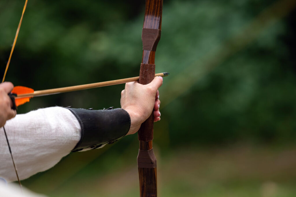 Person drawing a traditional wooden bow in the forest, demonstrating archery skills for survival and silent defense