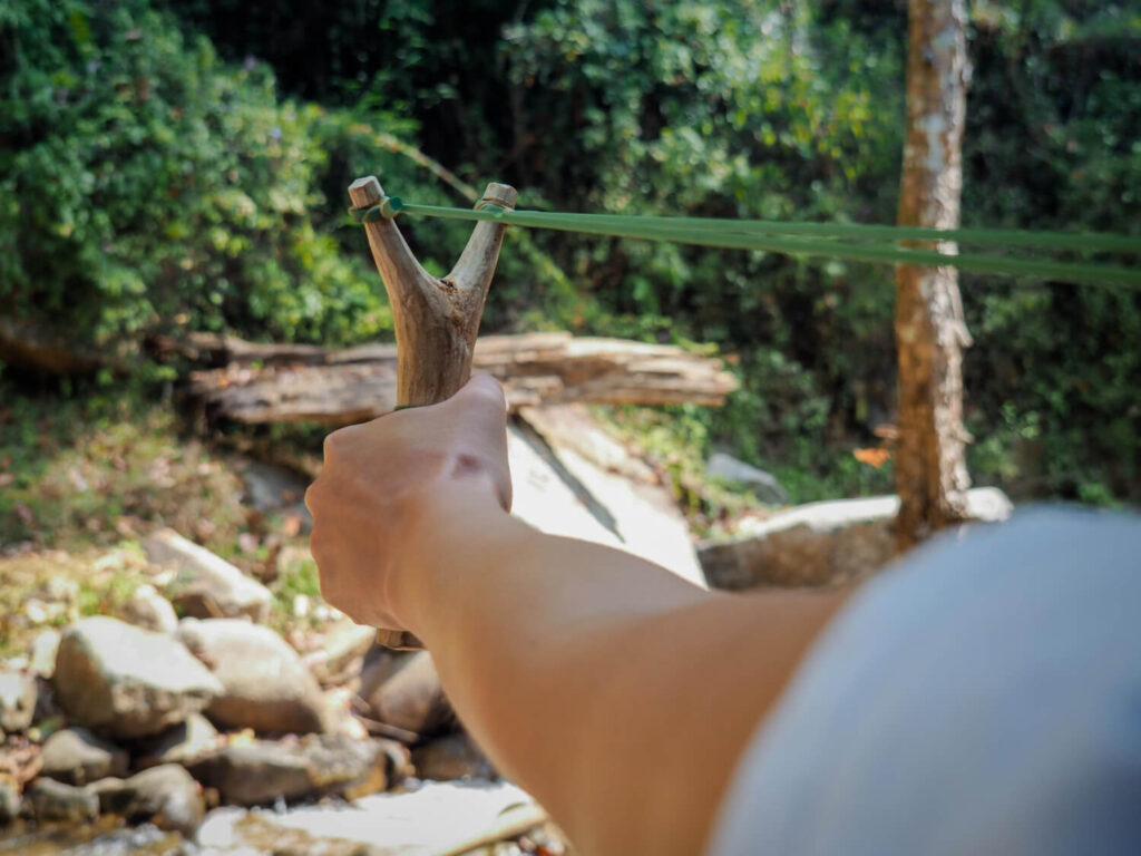 Hand aiming a homemade slingshot in a forest survival setting, representing improvised weapons for preppers