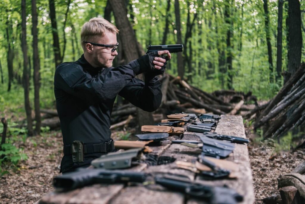 Prepper practicing handgun shooting at outdoor range with multiple weapons laid out on a wooden table