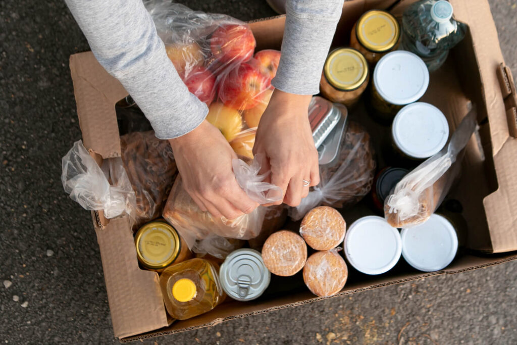 Hands organizing a food supply box with canned goods, oil, fruit, and shelf-stable nutrition