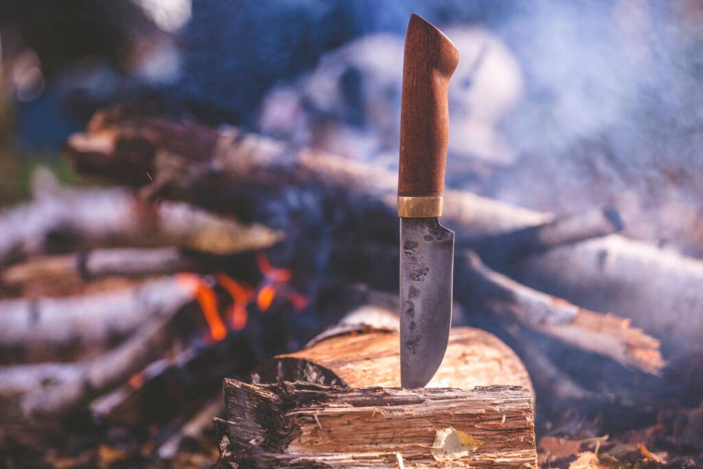 Survival knife stuck in a log near a campfire outdoors.