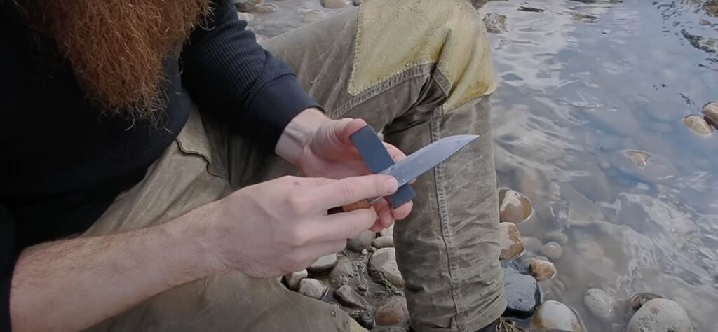 Hands sharpening a survival knife on a pocket whetstone outdoors by a river.