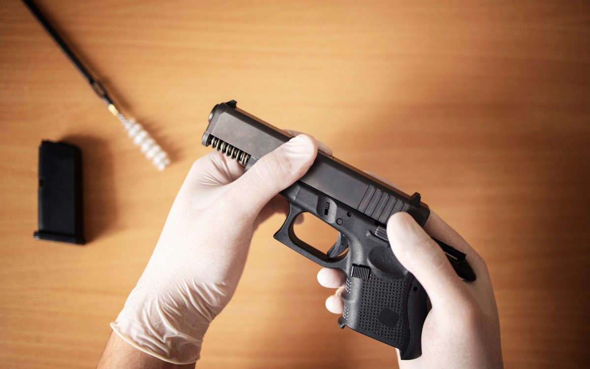 Person wearing gloves cleaning a handgun with a disassembled magazine and bore brush on a table.