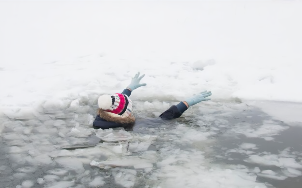 Person with raised arms struggling in icy water after falling through a frozen lake, wearing winter clothing and gloves.