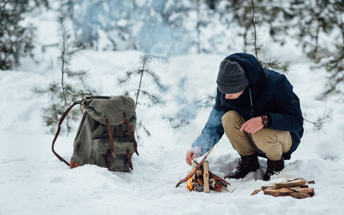 Man building a small fire in the snow with a survival backpack nearby, surrounded by a snowy forest.