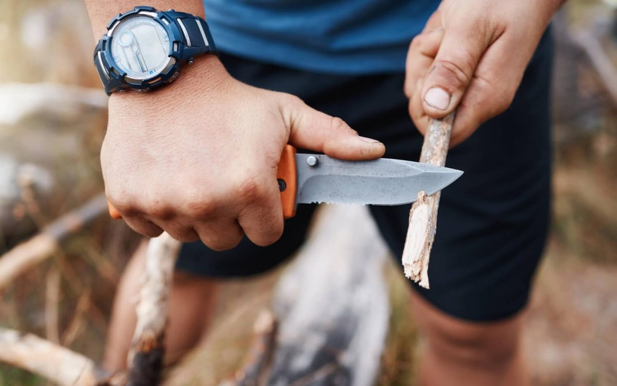 Person carving a stick with a survival knife outdoors, practicing essential knife skills.