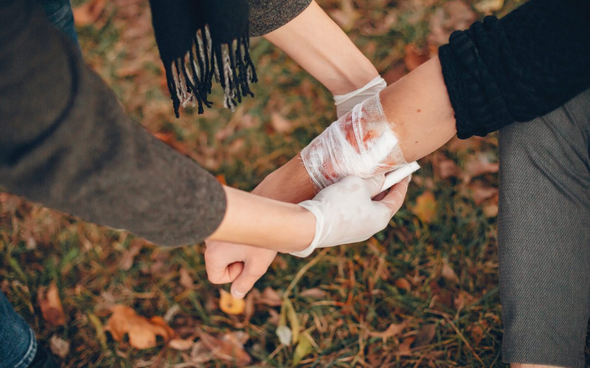 Person providing first-aid by wrapping a bandage around an injured arm outdoors, emphasizing the importance of basic first-aid in survival and outdoor scenarios.